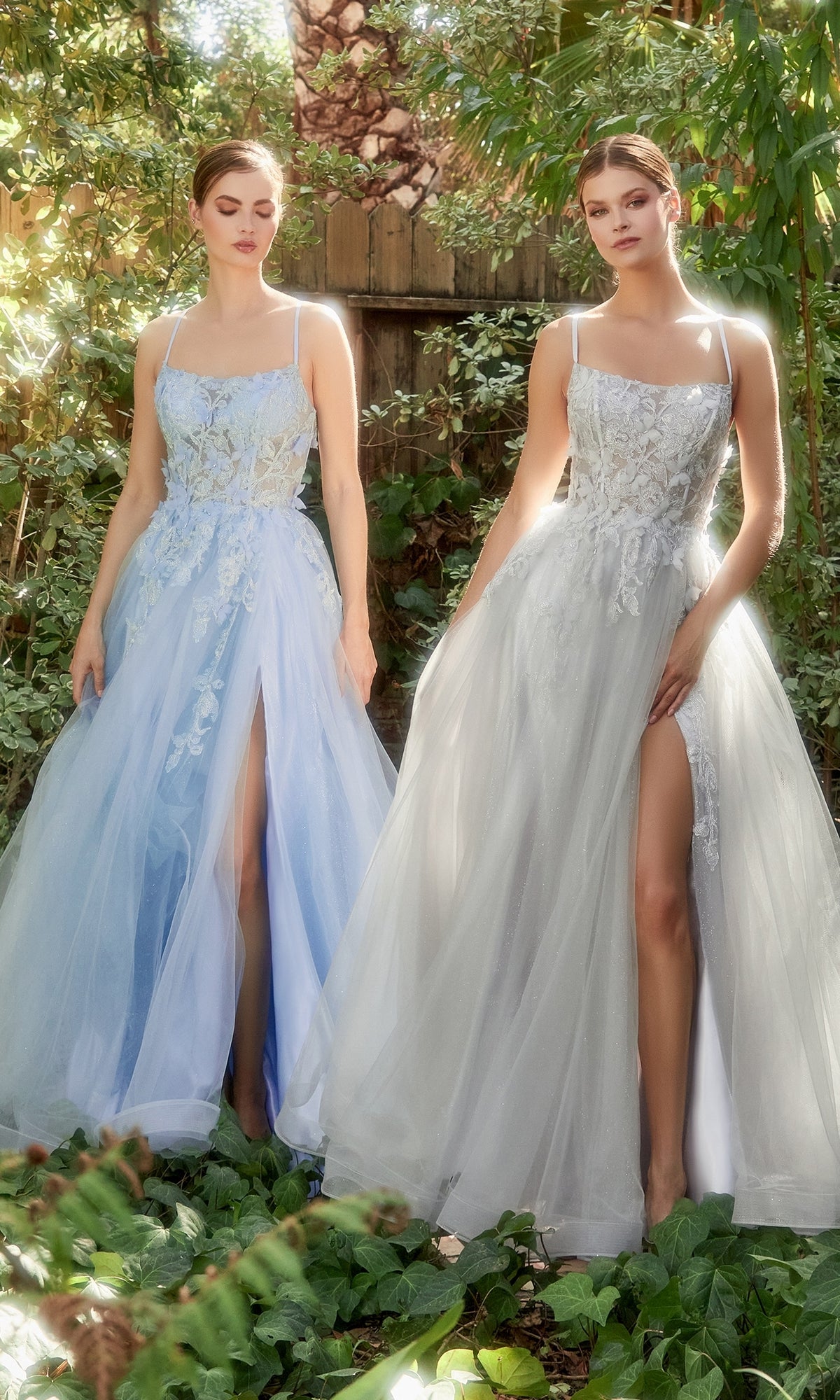 Two women stand outdoors in Butterfly Sheer-Bodice Prom Ball Gown A1141—sleeveless, floor-length dresses with floral embroidery and high slits, one light blue and one pale gray—amid lush greenery.